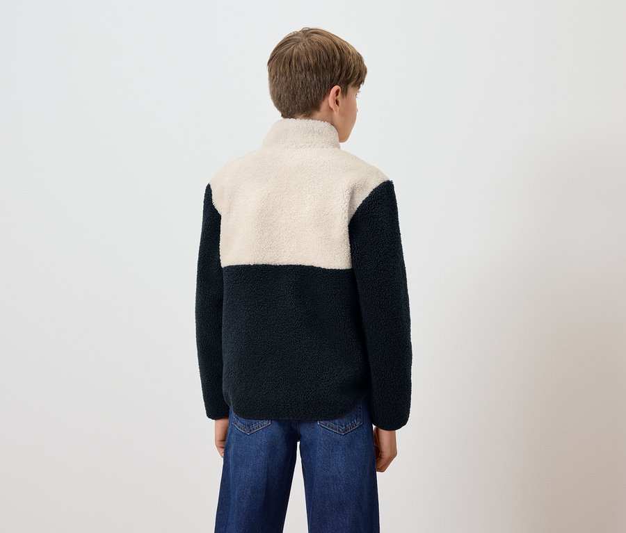 Un garçon, vu de dos, porte un jean bleu et une veste en polaire avec un haut crème et un bas bleu foncé.