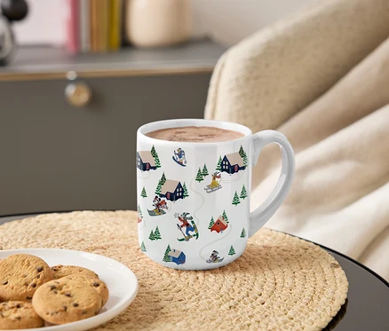 Une tasse avec un motif de Mickey et ses amis skiant dans les montagnes est posée sur une table à côté d'une assiette de biscuits.