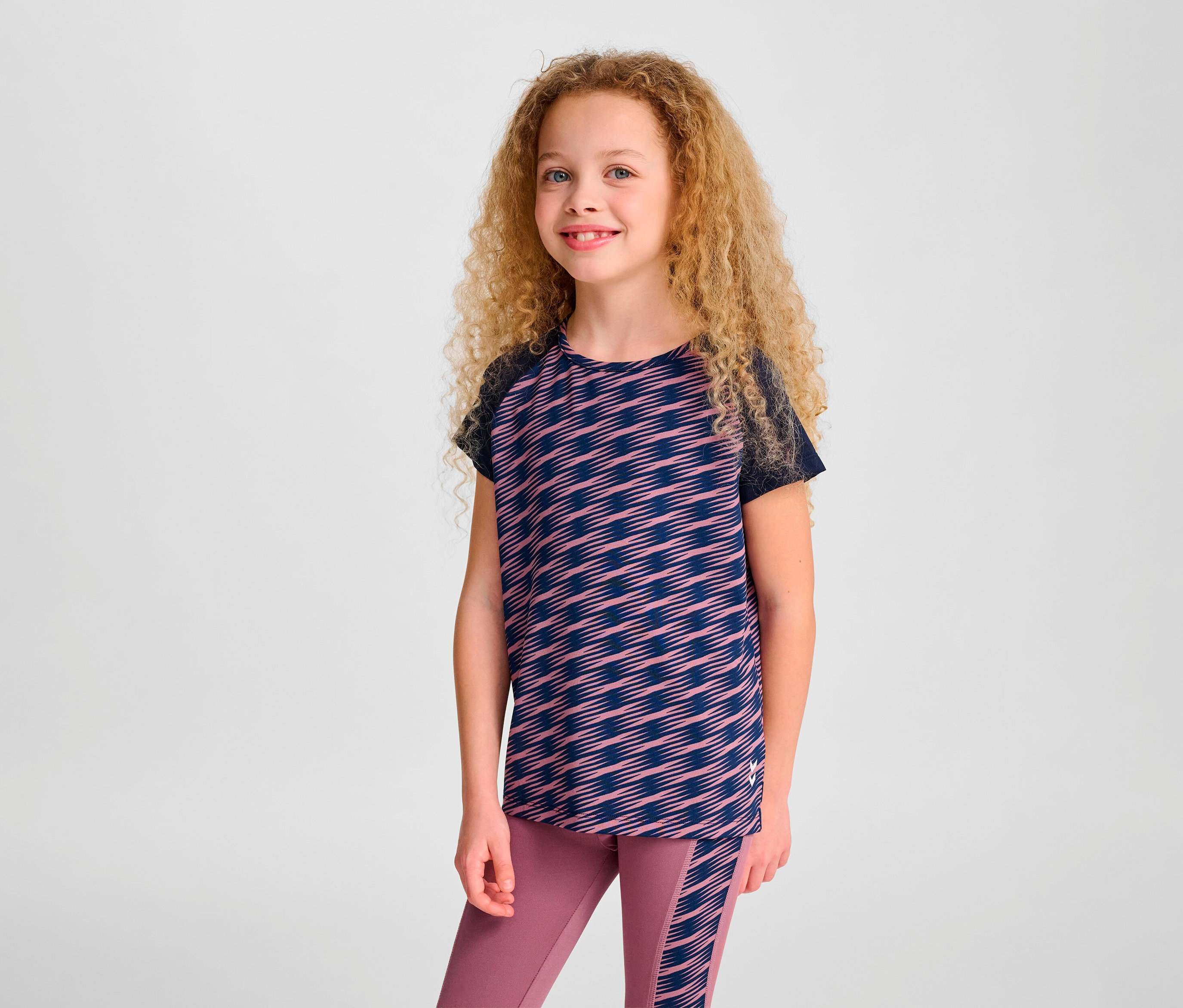 Une fille aux cheveux bouclés pose en T-shirt S/S HUMMEL HMLJR Mix et leggings.