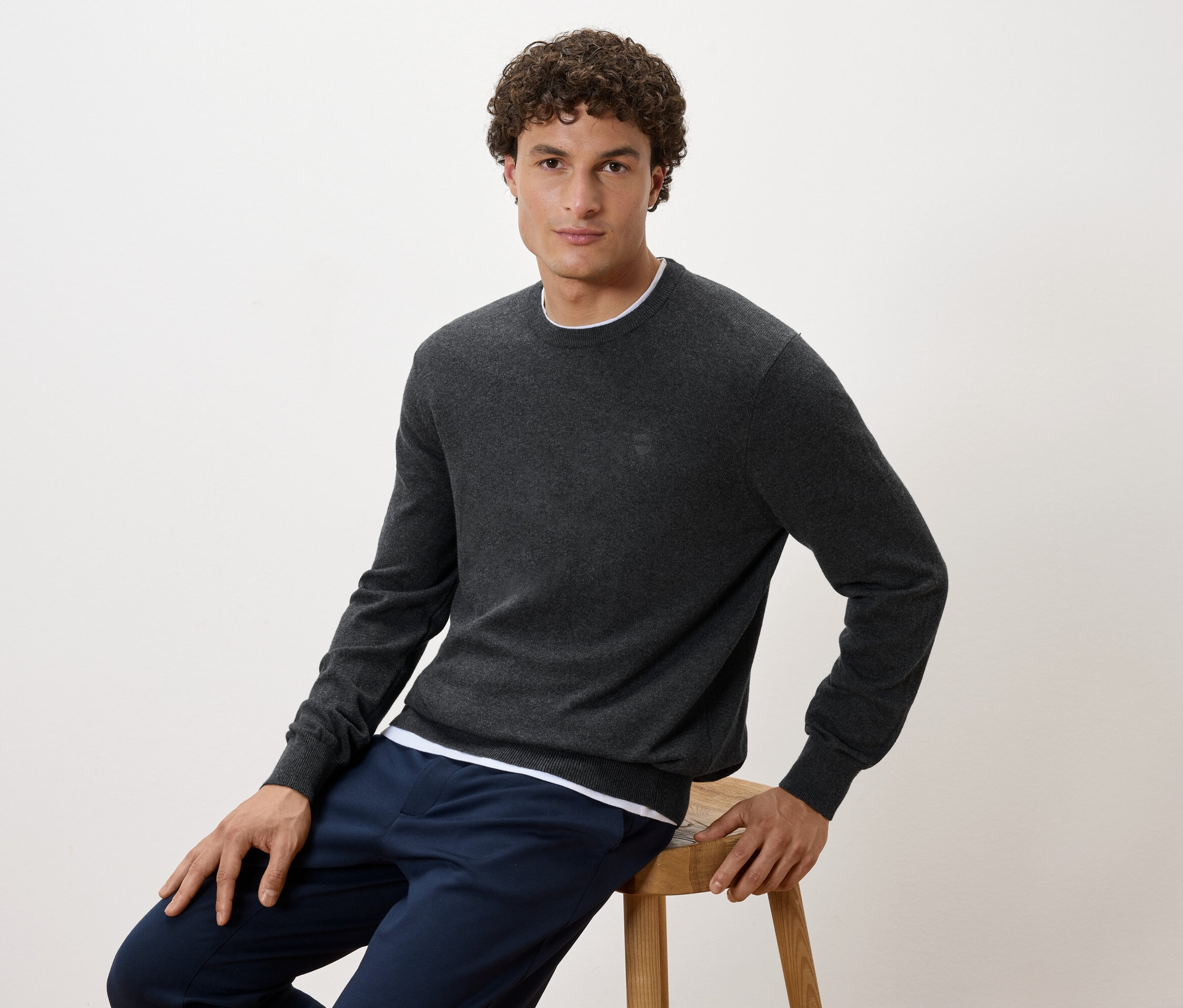 Un homme assis sur un tabouret porte un pull gris et un pantalon bleu marine.