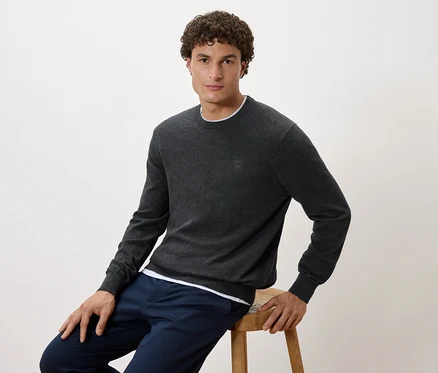 Un homme assis sur un tabouret porte un pull gris et un pantalon bleu marine.