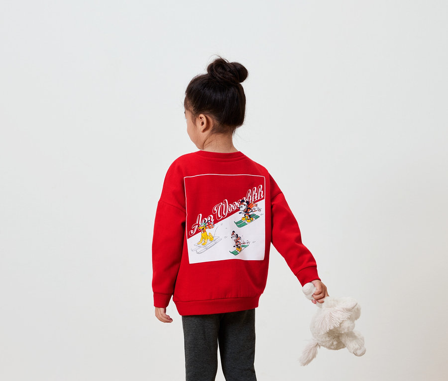 Une fille dos à la caméra porte un sweat-shirt rouge avec une image sur le dos et tient une peluche dans sa main.