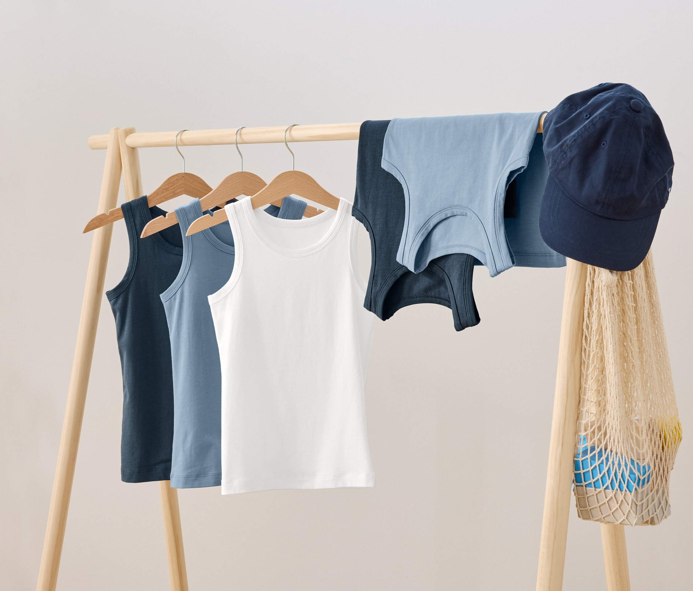 Sur un portant en bois, trois débardeurs sont suspendus à des cintres, à côté desquels sont posés deux autres débardeurs, une casquette bleue et un sac en filet.