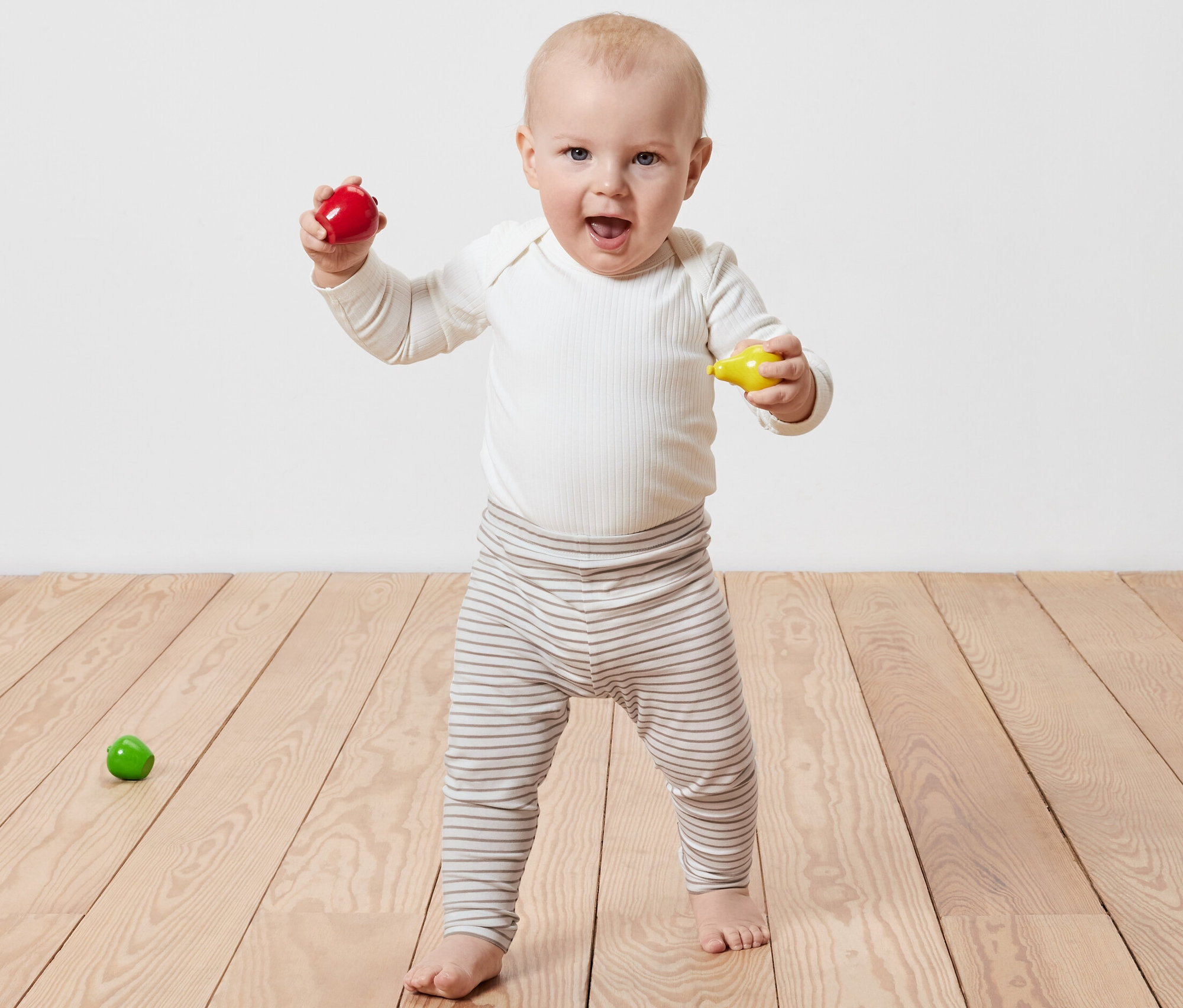 Un bébé souriant se tient sur un plancher en bois, tenant un jouet rouge et un jouet jaune dans ses mains. Il porte des 5 leggings enfant.