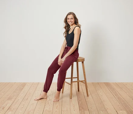 Une femme souriante est assise sur un tabouret en bois, portant un débardeur noir et un pantalon de jogging bordeaux.