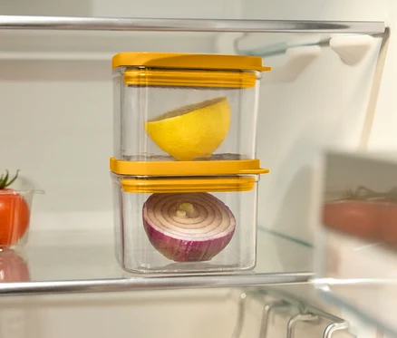 Dans le réfrigérateur, deux récipients transparents avec des couvercles jaunes sont empilés l'un sur l'autre. Le récipient supérieur contient un demi-citron et le récipient inférieur un demi-oignon rouge. À gauche, il y a un récipient en plastique avec des tomates.