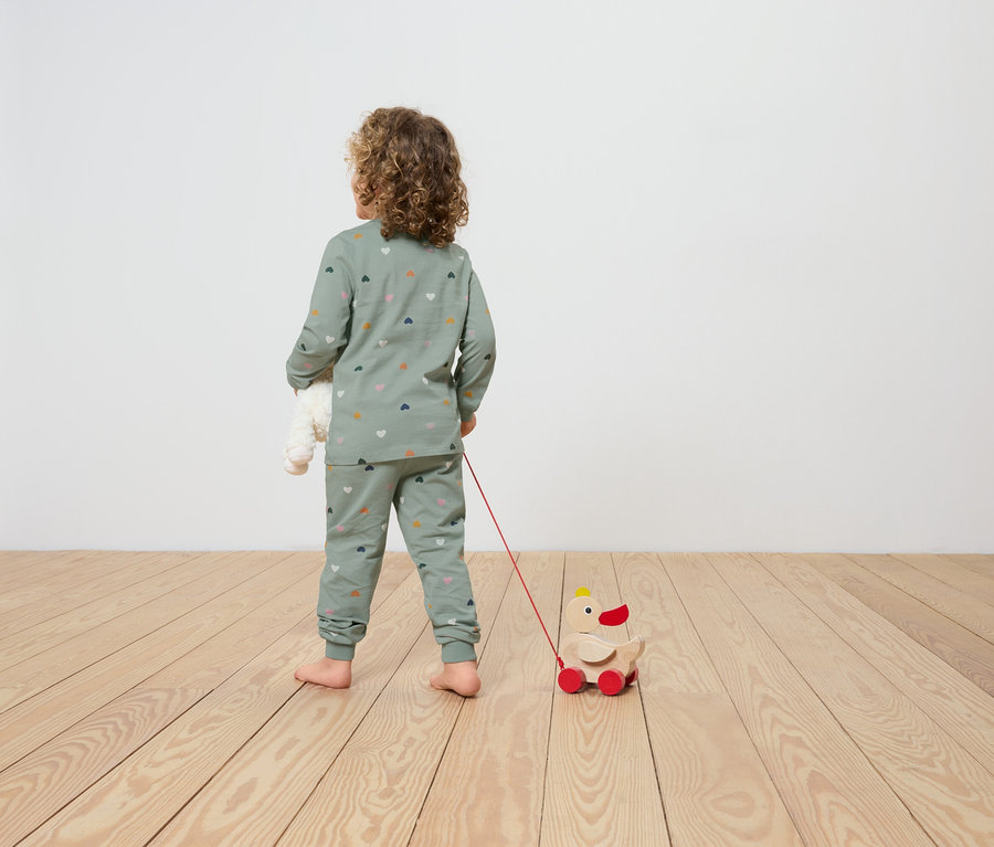 Un enfant en pyjama imprimé avec des coeurs tire un jouet de canard en bois sur roues et tient un ours en peluche dans ses bras.