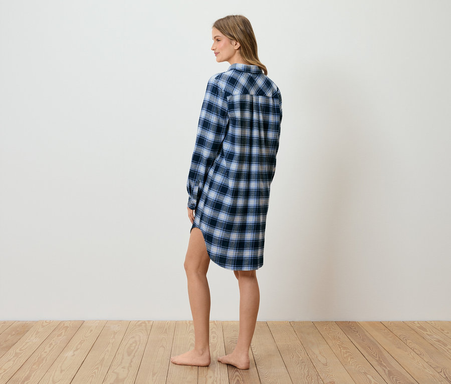 Femme en chemise de nuit à carreaux bleue, debout de profil sur un plancher en bois devant un mur blanc.