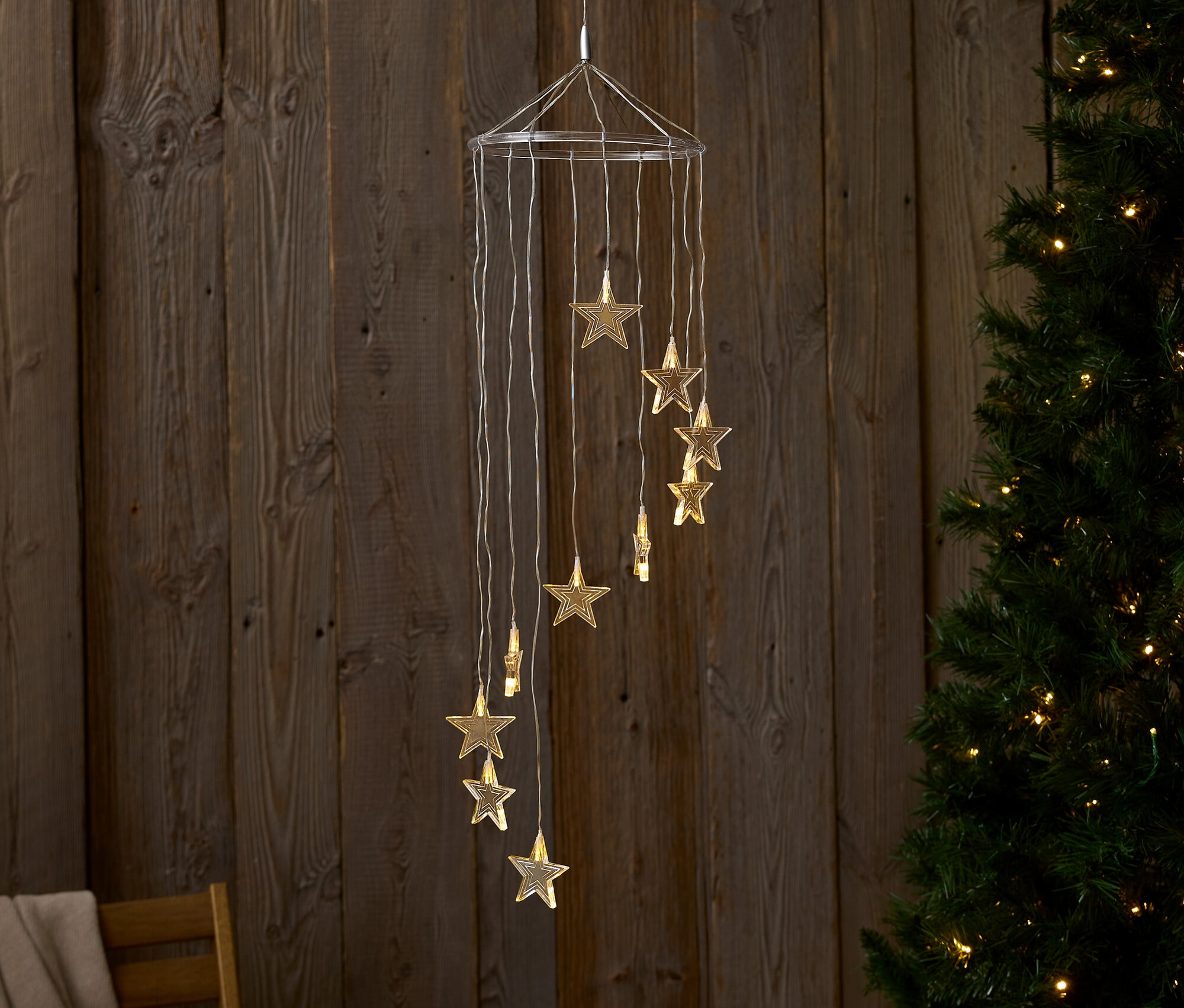 Une décoration suspendue avec des lumières en forme d'étoiles devant un mur en bois et un arbre de Noël.
