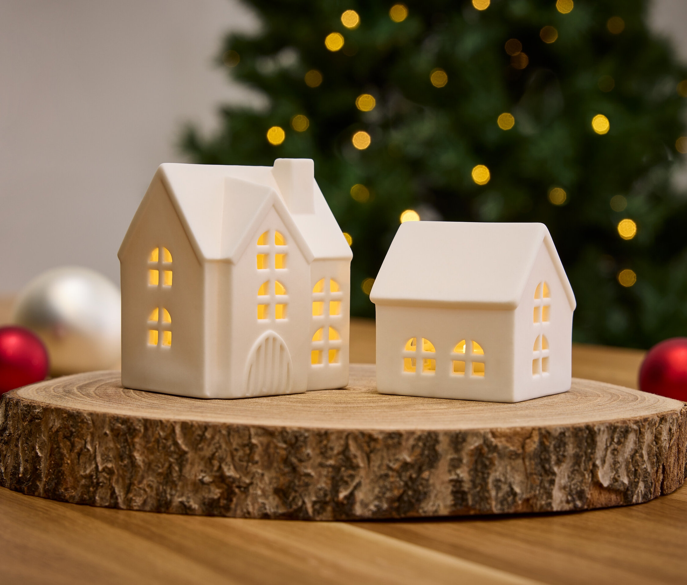 Deux petites maisons blanches sur une base en bois avec des décorations de Noël et un arbre en arrière-plan.