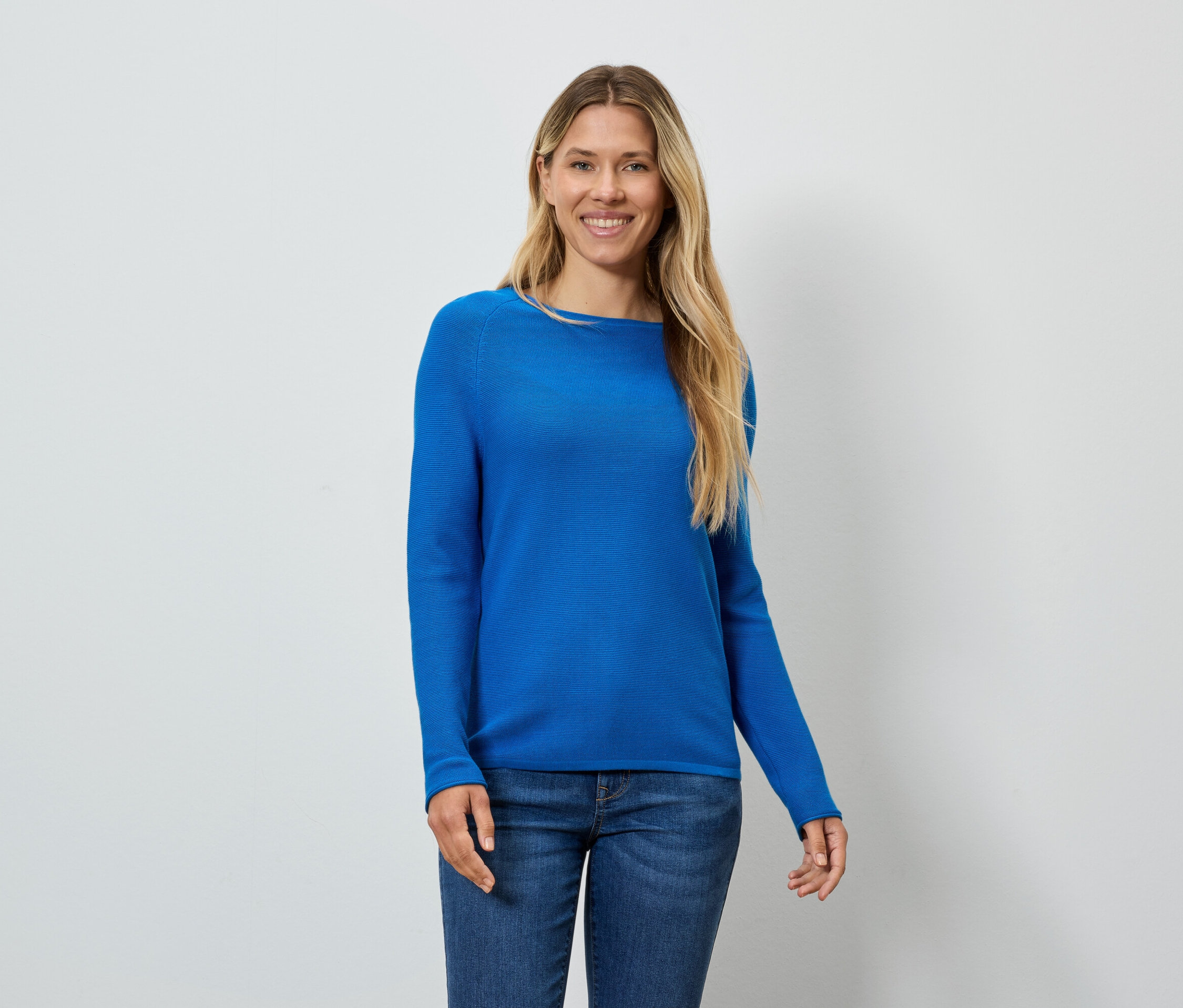 Une femme blonde souriante porte un pull bleu et un jean.