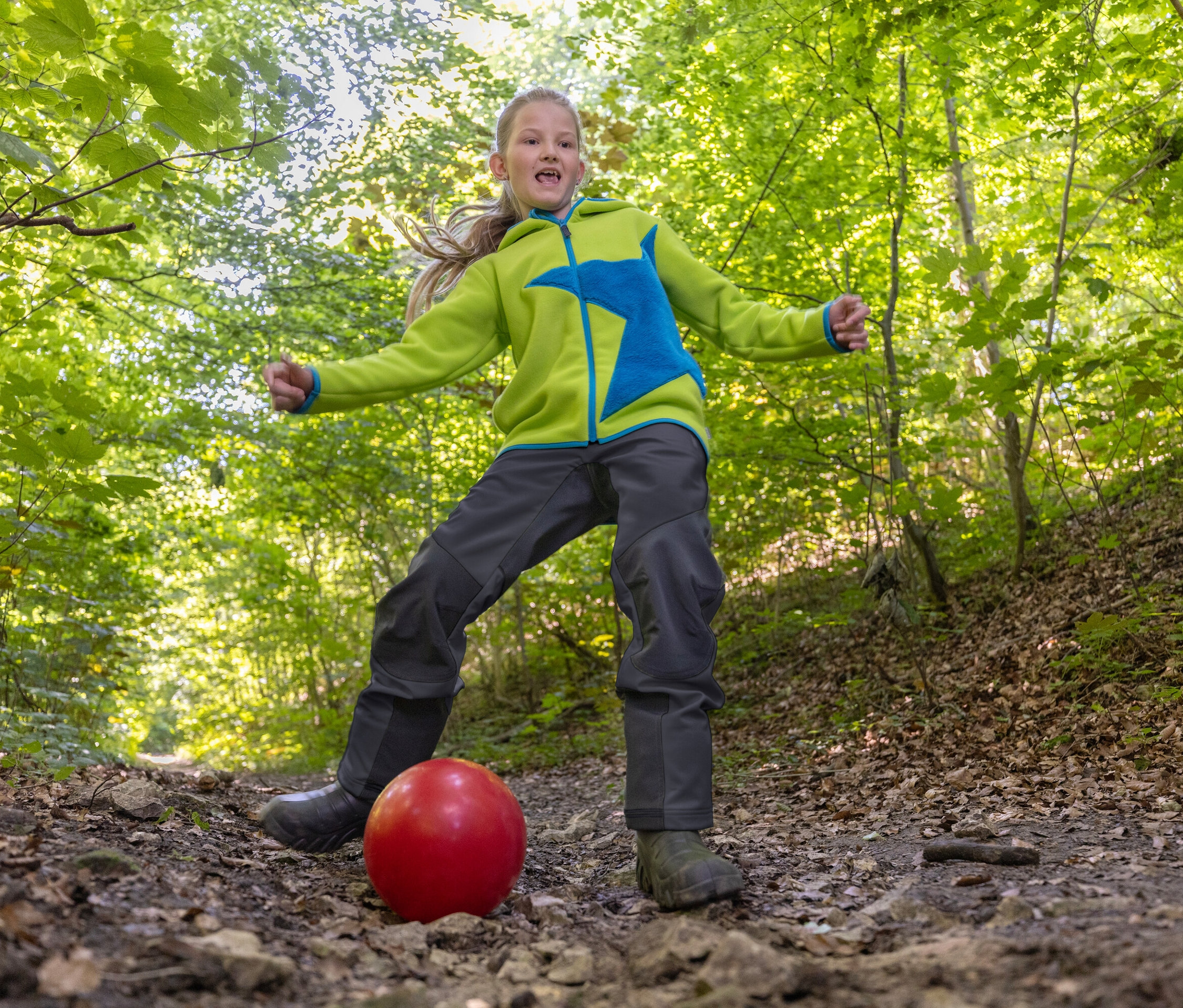 Une fille tape dans un ballon rouge sur un chemin forestier. La fille porte une veste verte avec une étoile bleue et un pantalon foncé.