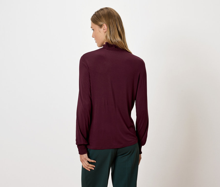 Femme portant un chemisier bordeaux à manches longues et à col roulé et un pantalon vert, vue de dos.