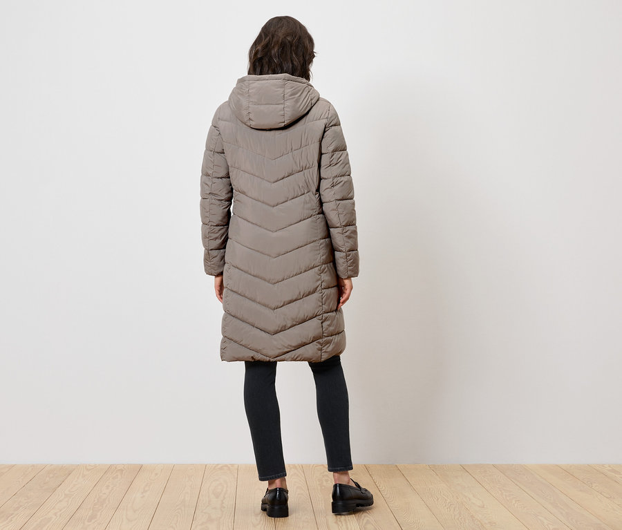 Une femme vue de dos porte un long manteau matelassé à capuche, un pantalon noir et des chaussures noires.