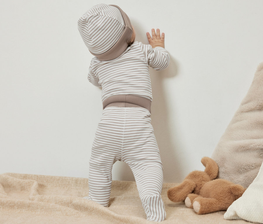 Un bébé dans un ensemble pour bébé 3 pièces debout près d'un mur.