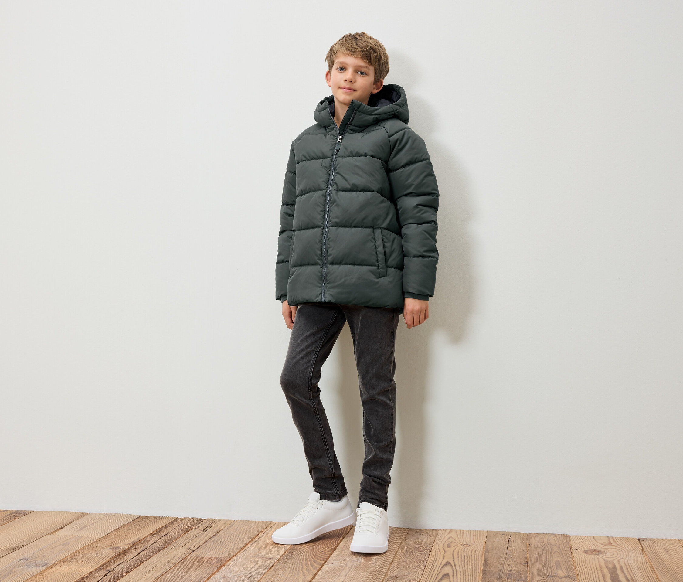 Un garçon debout portant une doudoune vert foncé, un jean gris et des baskets blanches.