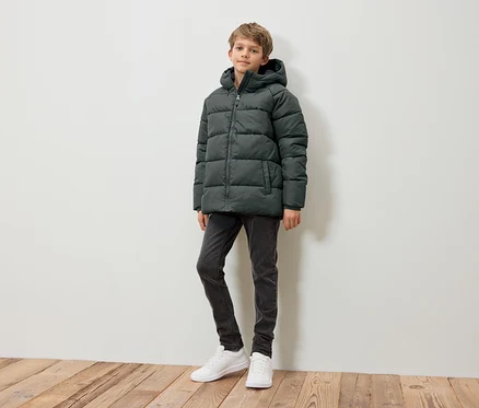 Un garçon debout portant une doudoune vert foncé, un jean gris et des baskets blanches.