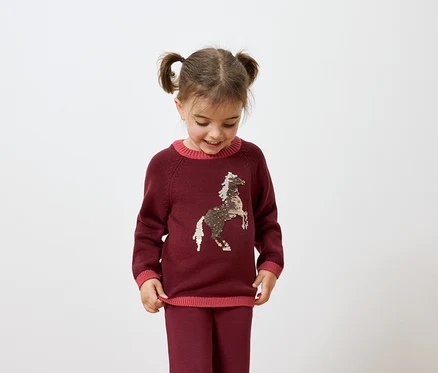 Une fille avec des couettes porte un pull bordeaux avec un cheval et un pantalon bordeaux.