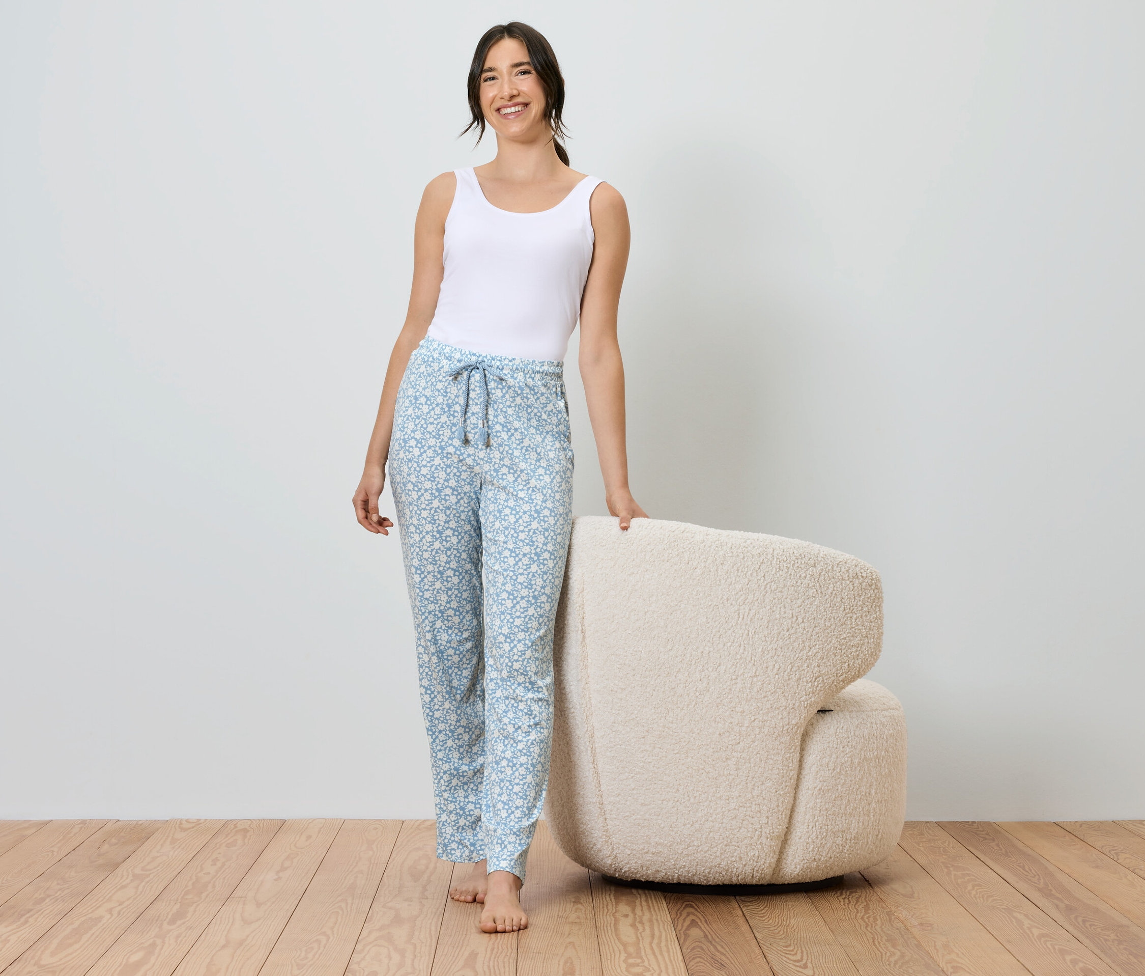 Une femme en débardeur blanc et pantalon bleu à motifs se tient à côté d'un fauteuil crème.