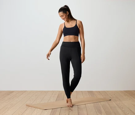 Une femme debout sur un tapis de yoga portant un soutien-gorge de sport noir et un legging noir.