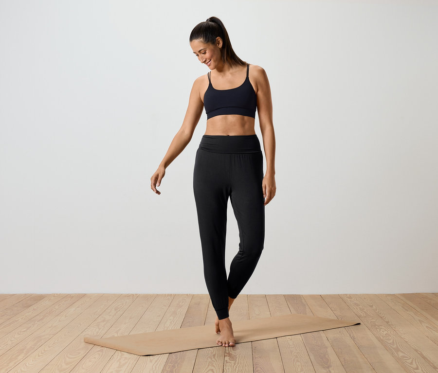Une femme debout sur un tapis de yoga portant un soutien-gorge de sport noir et un legging noir.