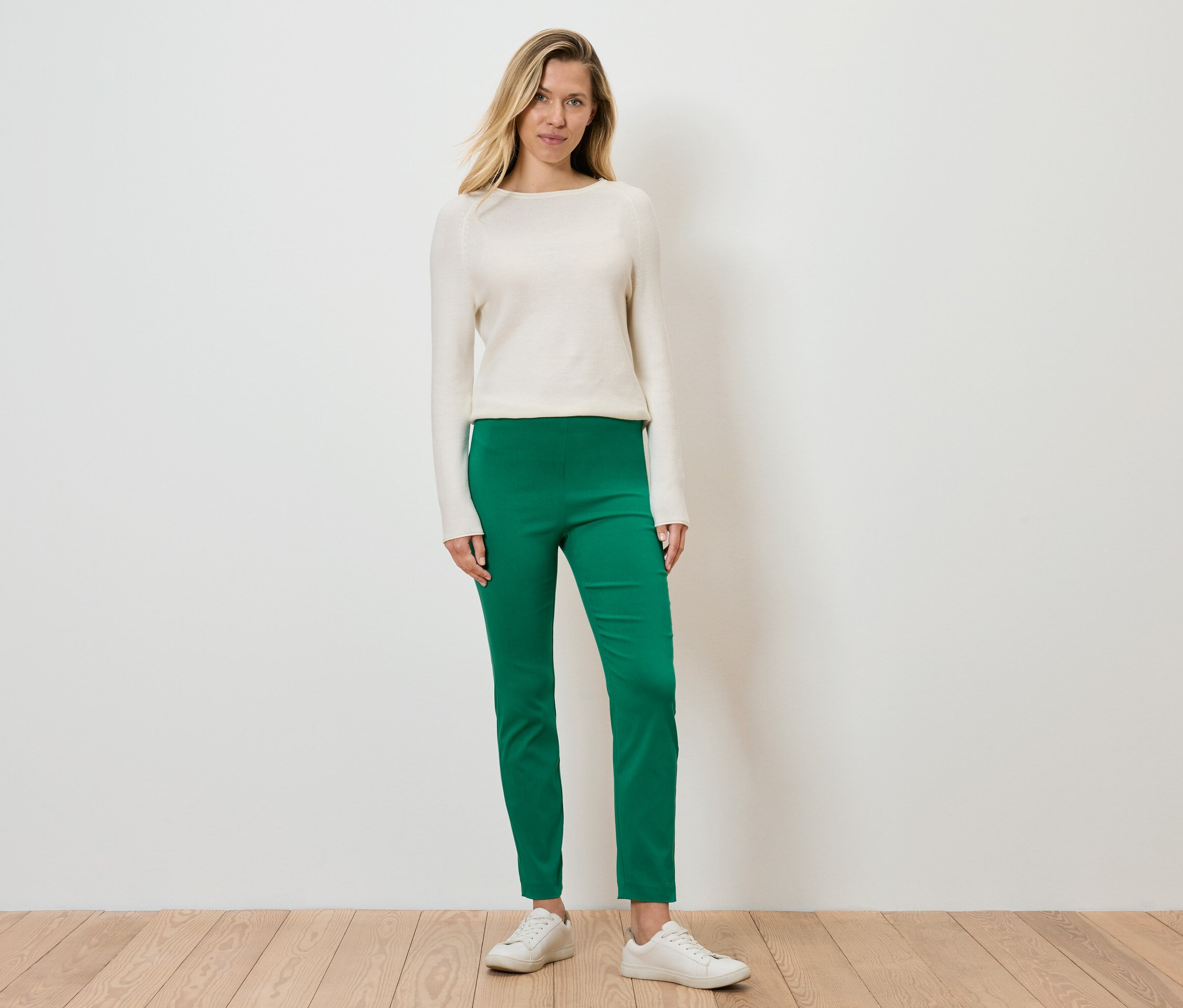Une femme debout porte un pull couleur crème, un pantalon vert et des baskets blanches.