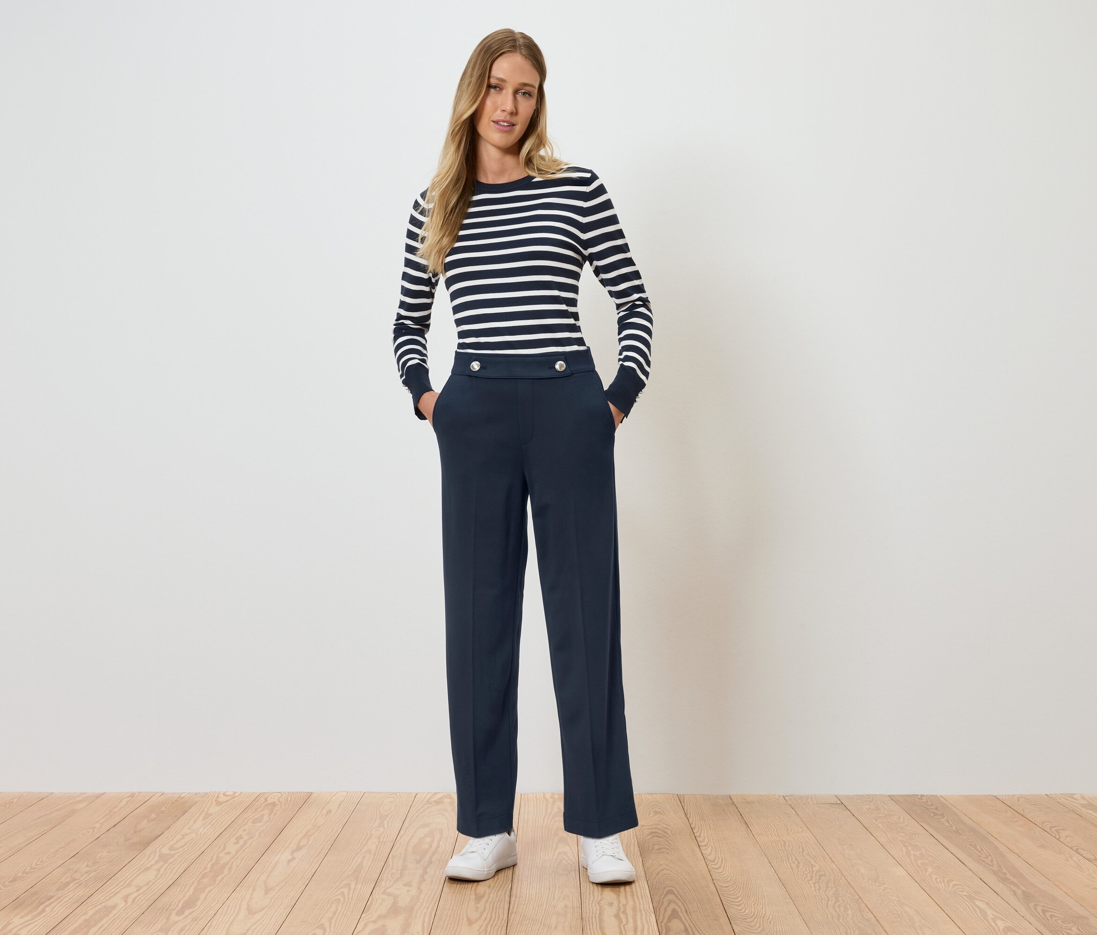 Une femme debout, les mains dans les poches. Elle porte un T-shirt rayé à manches longues et un pantalon bleu marine.