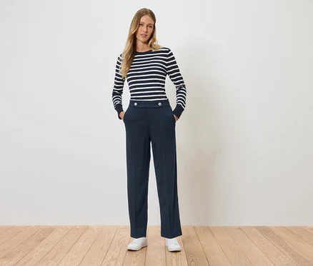 Une femme debout, les mains dans les poches. Elle porte un T-shirt rayé à manches longues et un pantalon bleu marine.