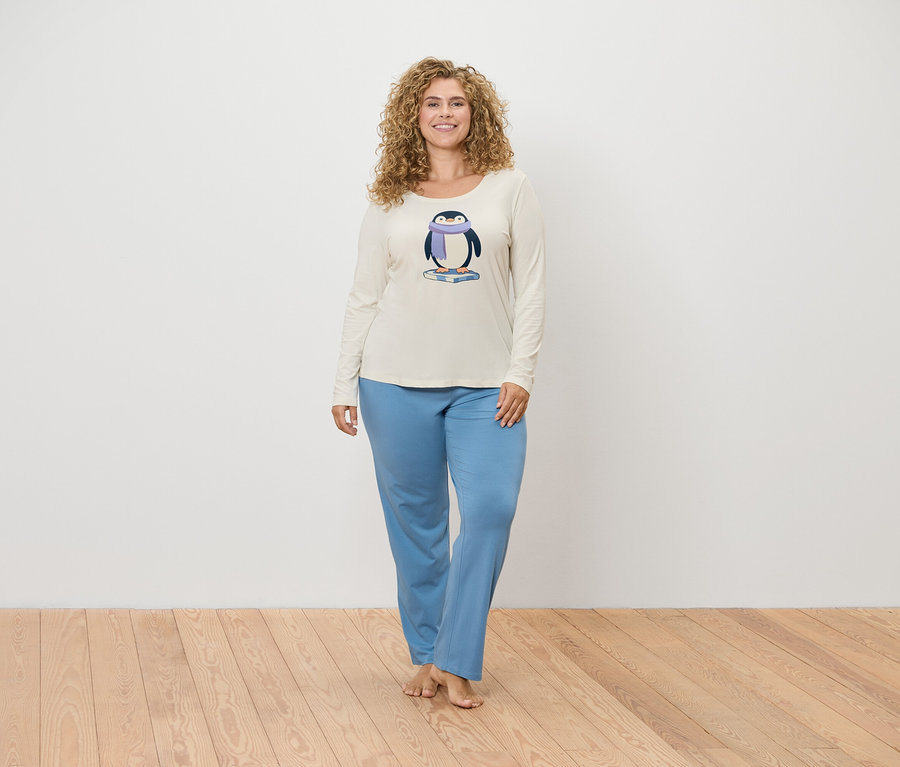 Femme posant dans un haut de pyjama crème avec un imprimé de pingouin et un pantalon de pyjama bleu.