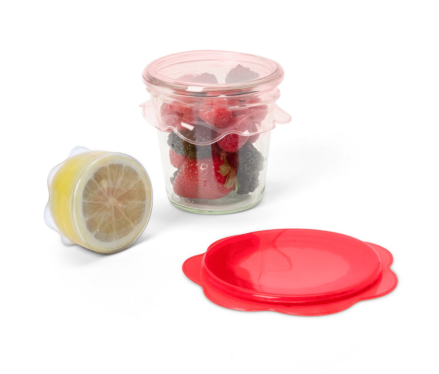 Un bocal de fruits recouvert d'un des 3 couvercles en silicone, à côté d'un demi-citron recouvert du même couvercle et un couvercle rouge.