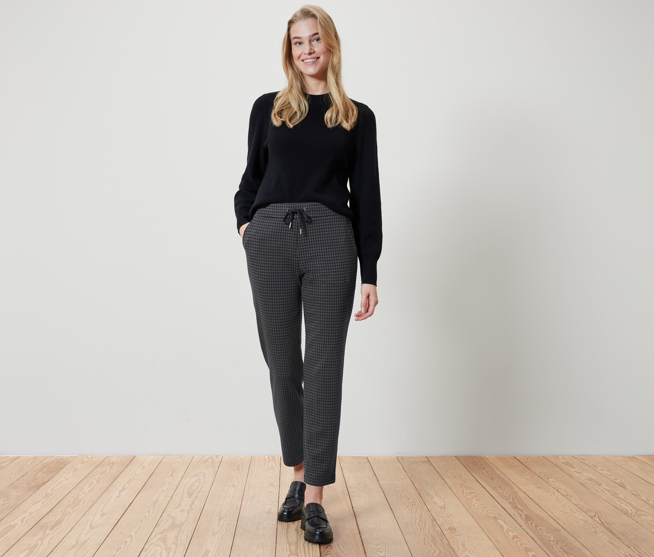 Une femme se tient debout, les mains dans les poches d'un pantalon à carreaux. Elle porte un pull noir et des chaussures noires.