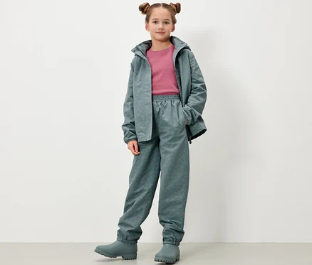 Une fille pose, portant un pantalon de pluie pour enfants avec des éléments réfléchissants, une veste et des bottes en caoutchouc gris-vert.