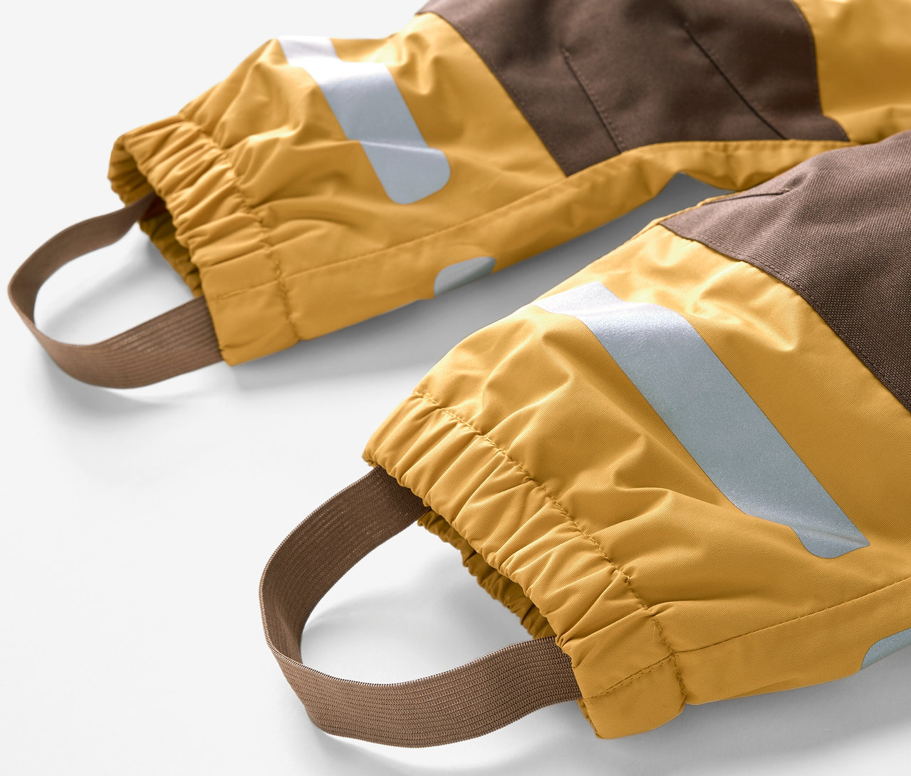 Gros plan sur les chevilles d'une combinaison de pluie pour enfant, avec des blocs de couleurs marron et jaune et des élastiques de maintien au niveau des pieds.