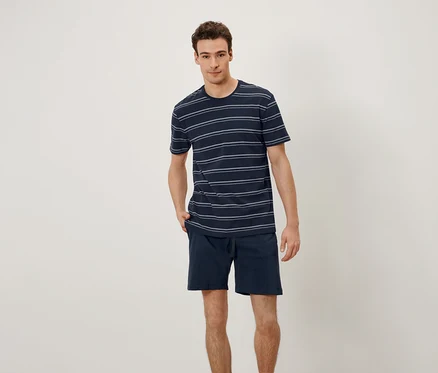 Un homme se tient debout, les mains dans les poches. Il porte un t-shirt à rayures horizontales et un short.