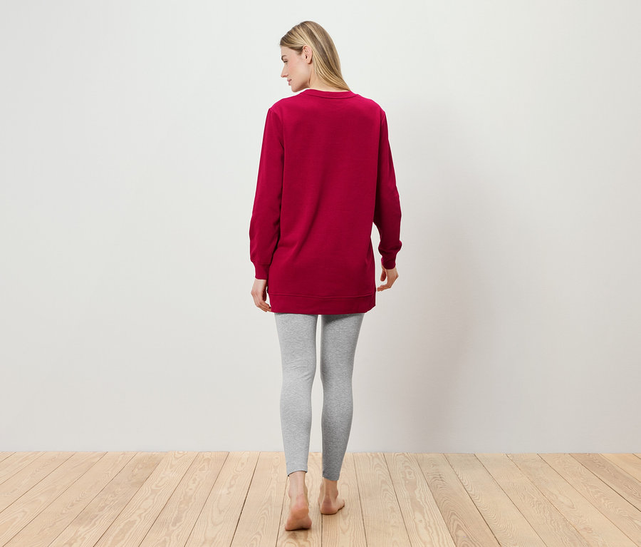 Une femme se tient dos à la caméra portant une tunique bordeaux et des leggings gris.