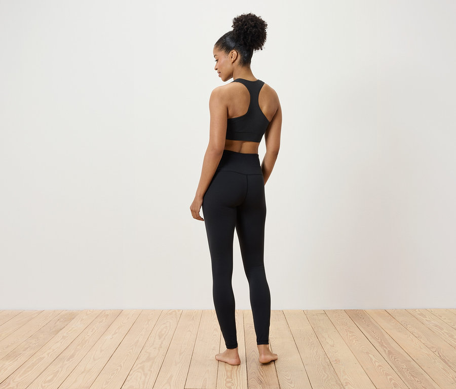 Une femme en soutien-gorge de sport noir et leggings debout sur un plancher en bois, dos à la caméra.