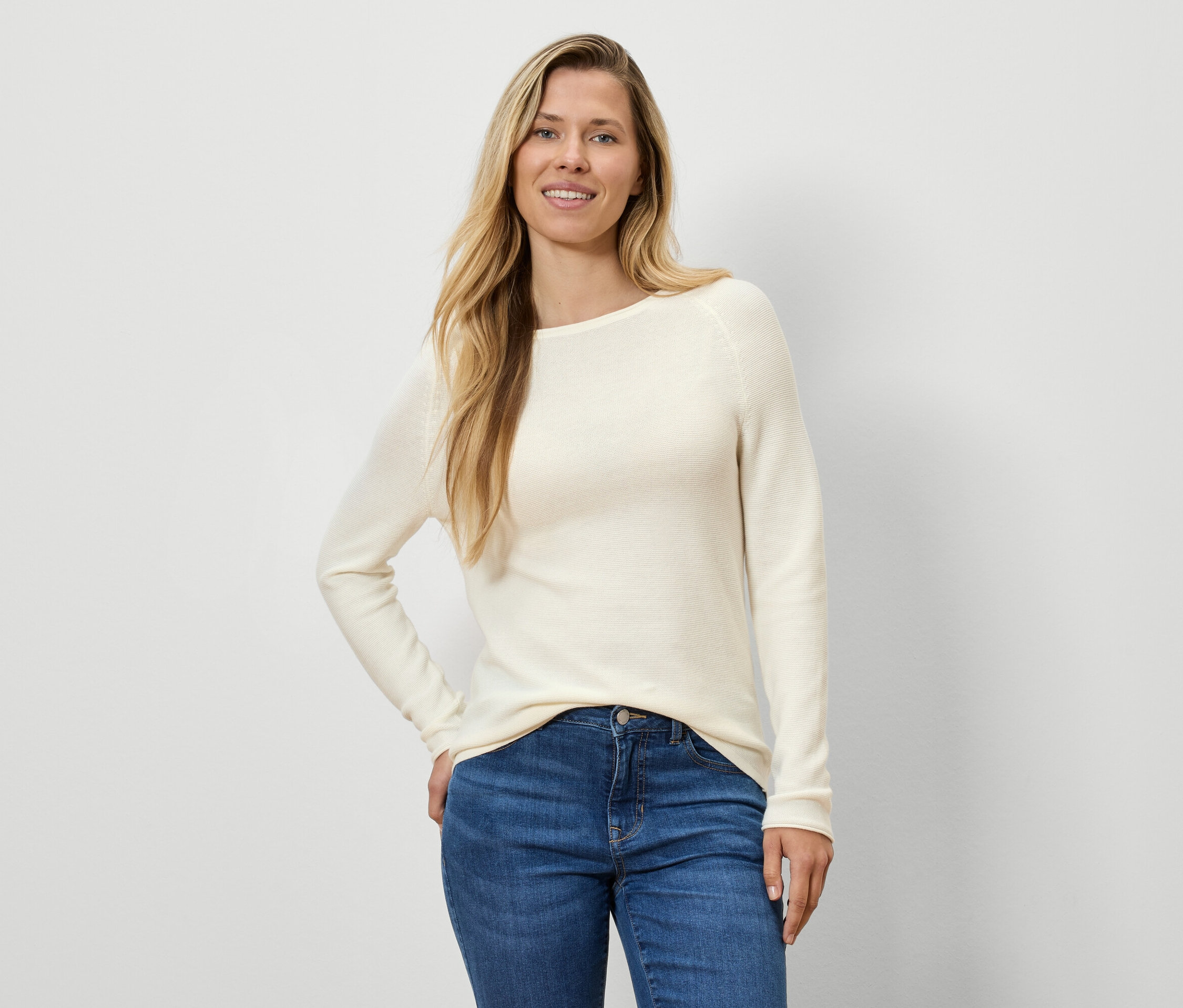 Une femme aux cheveux blonds pose dans un pull crème et un jean bleu.