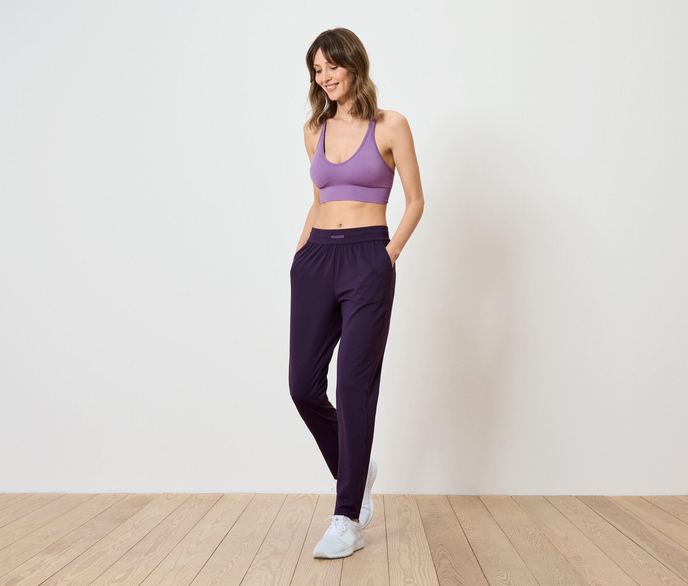 Une femme vêtue d'un soutien-gorge de sport lilas et d'un pantalon violet foncé se tient sur un plancher en bois.