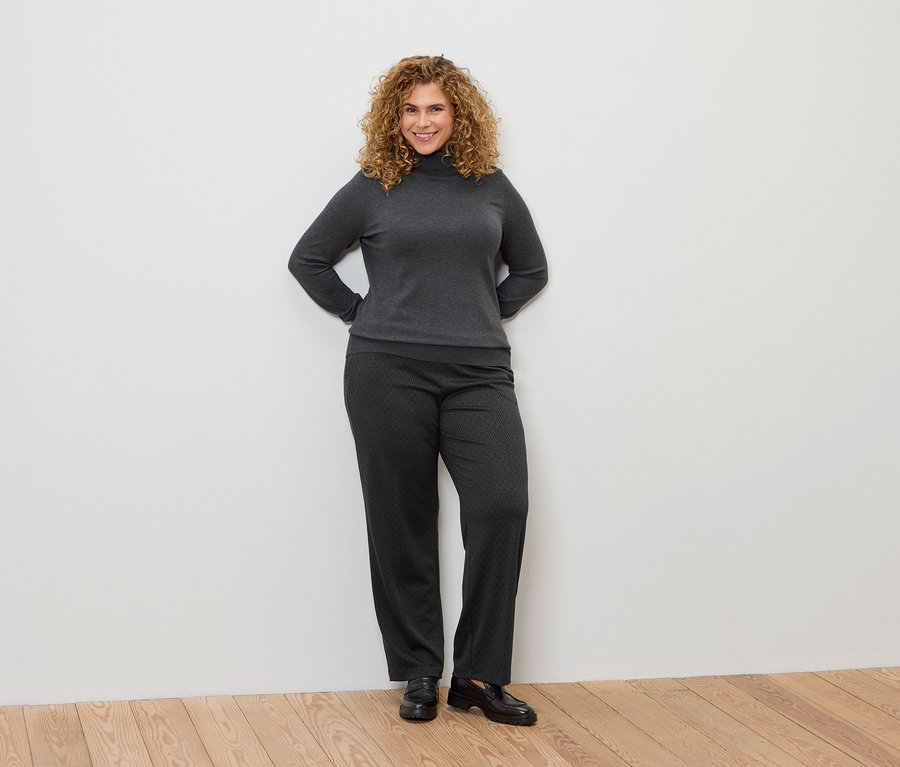 Une femme aux cheveux bouclés pose en col roulé gris et pantalon à motifs.