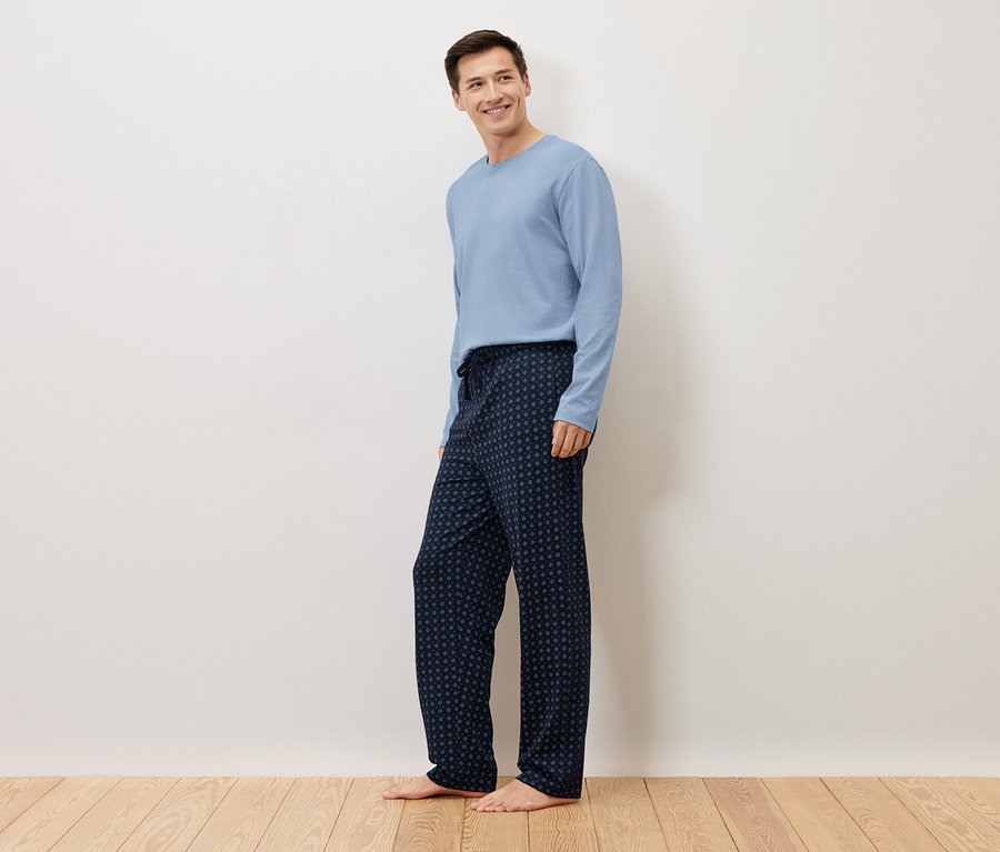 Un homme se tient pieds nus devant un mur blanc, portant un pull bleu clair et un pantalon de pyjama bleu marine à pois.