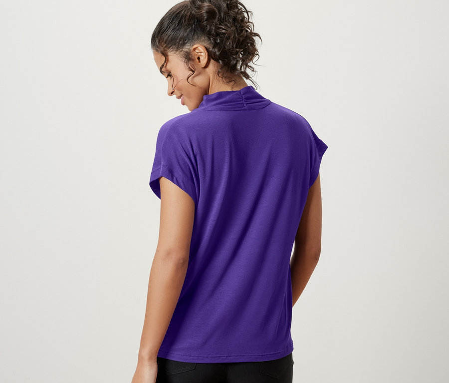 Une femme, vue de dos, porte une blouse violette avec un col en V et un short noir.