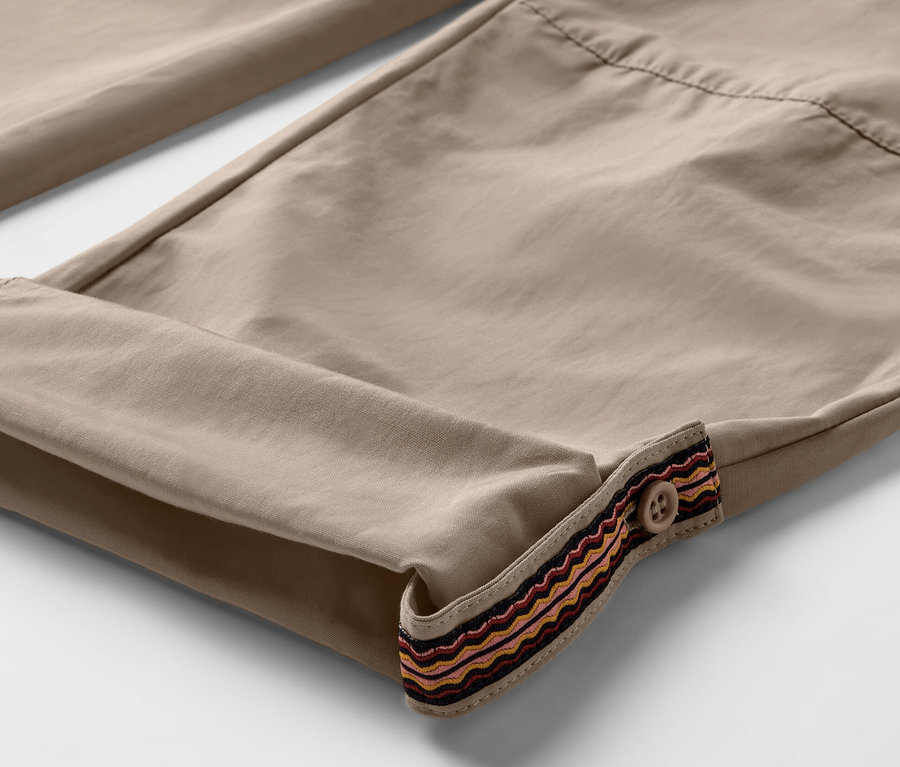 Gros plan sur un pantalon d’extérieur beige avec des bas de jambes retroussés et une bordure décorative avec un bouton. Le pantalon offre une protection anti-moustiques.