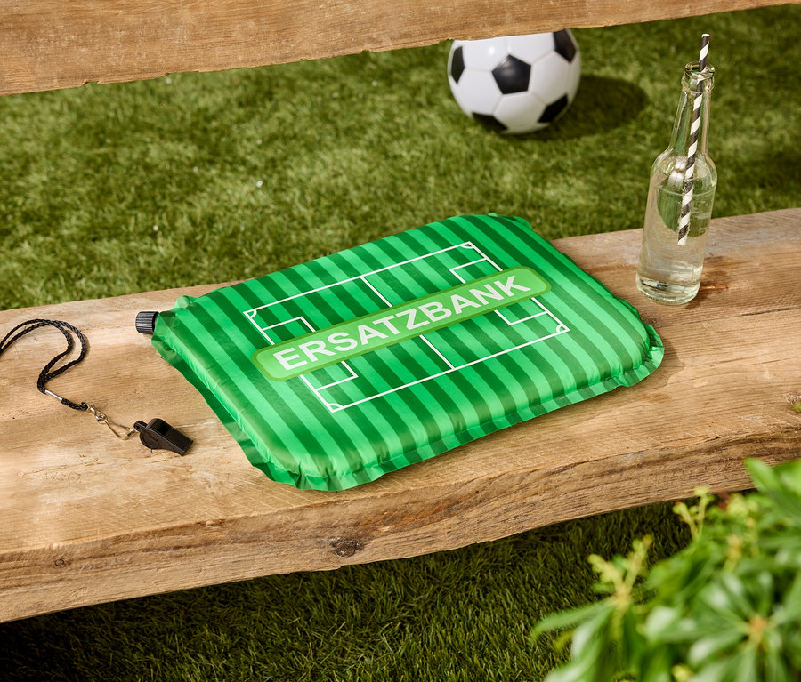 Sur un banc en bois se trouvent un coussin vert avec l'inscription 'ERSATZBANK', un sifflet, une bouteille d'eau et un ballon de football.