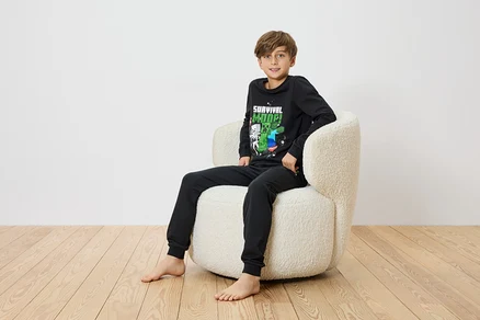 Un garçon est assis pieds nus dans un fauteuil crème et porte un pyjama noir Minecraft.