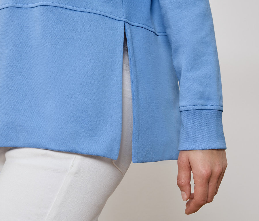 Une personne porte un sweat bleu clair avec une fente latérale et un pantalon blanc.