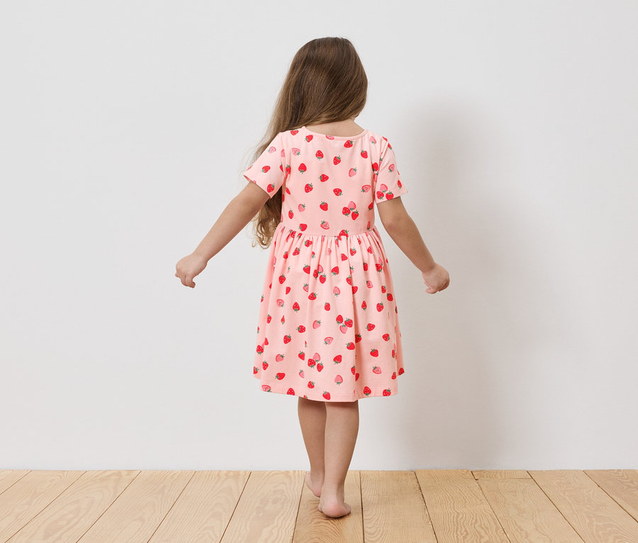 Fille vue de dos dans une robe rose pâle à motifs de fraises, debout sur un plancher en bois.
