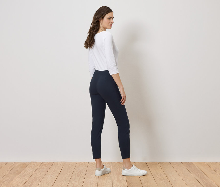 Femme portant un haut blanc et des leggings bleu marine posant devant un mur blanc sur un plancher en bois.