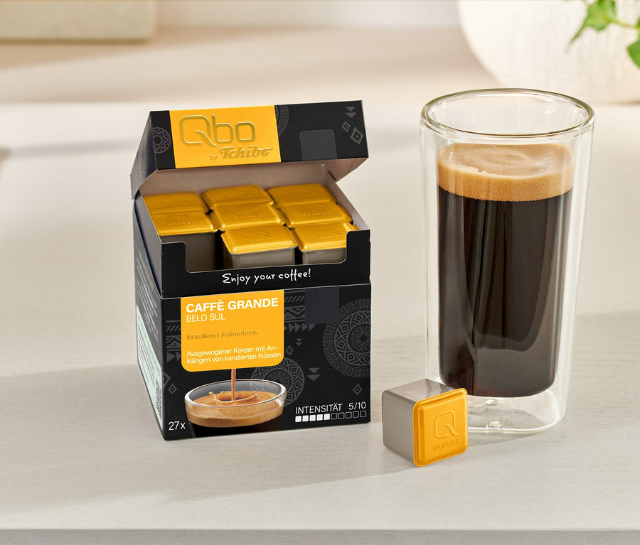 Une boîte ouverte de capsules Qbo Caffè Grande BELO SUL, une capsule individuelle et un verre de café sont posés sur une table.