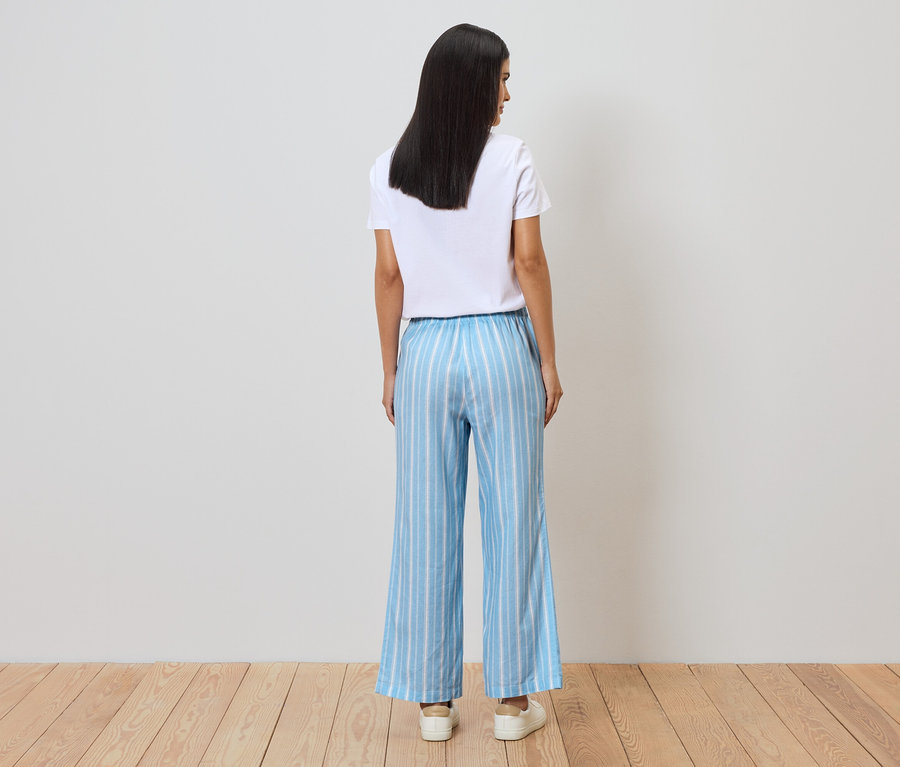 Vue de dos d'une femme portant un t-shirt blanc et un pantalon large rayé bleu et blanc devant un mur blanc.