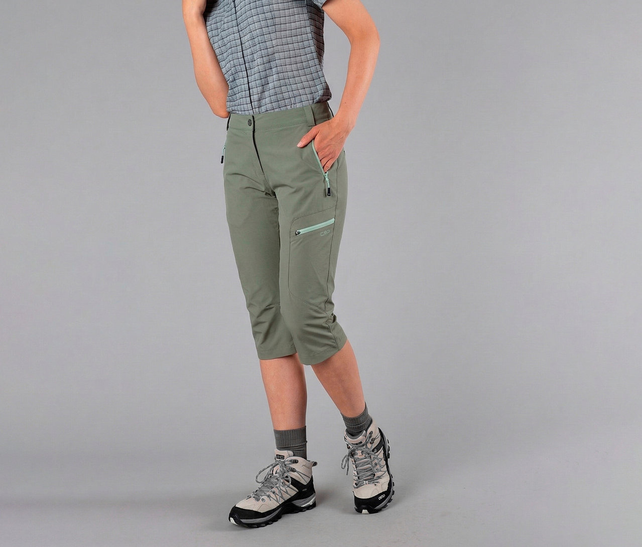 Femme portant une chemise à carreaux et un pantalon capri pour femme CPM en nylon stretch vert olive, les mains dans les poches et des chaussures de randonnée.
